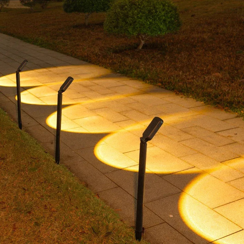 Four black LED solar landscape spotlights illuminate a stone pathway with warm light.