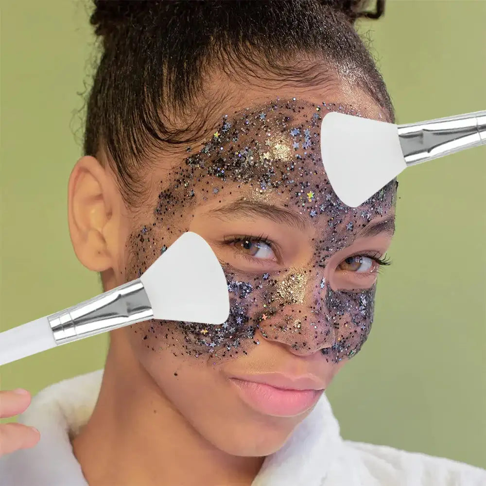 Two white, fan-shaped silicone face mask brushes with silver ferrules applying glittery mud mask to a person's face.