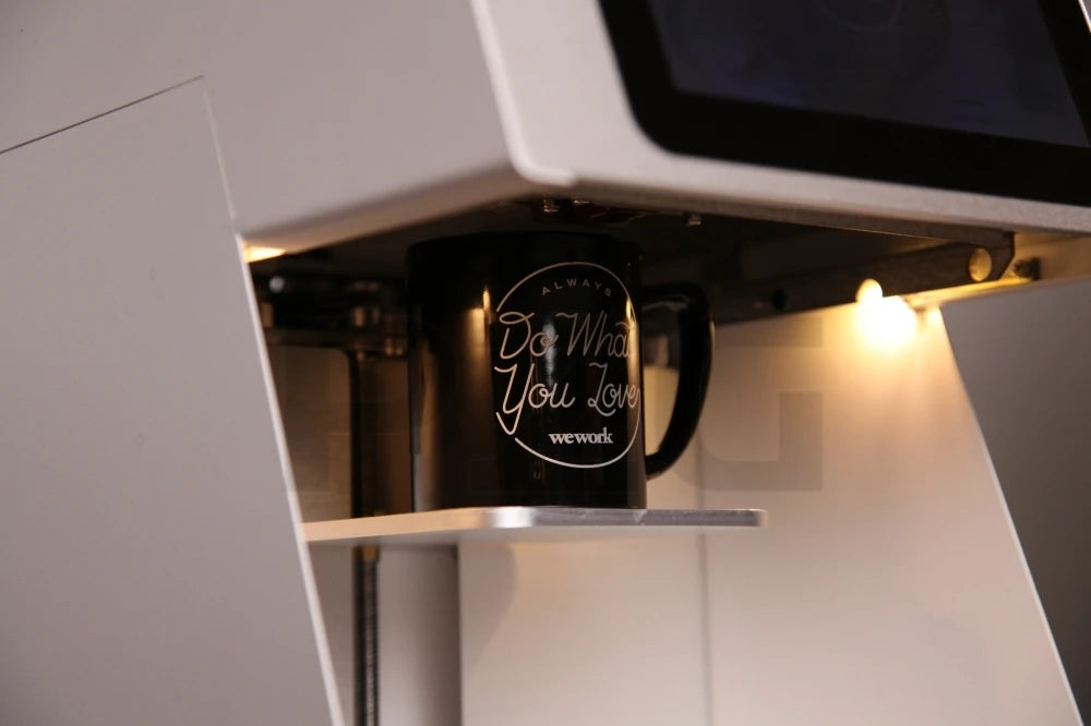 Black WeWork mug inside a modern white automated coffee machine with a digital touchscreen display.