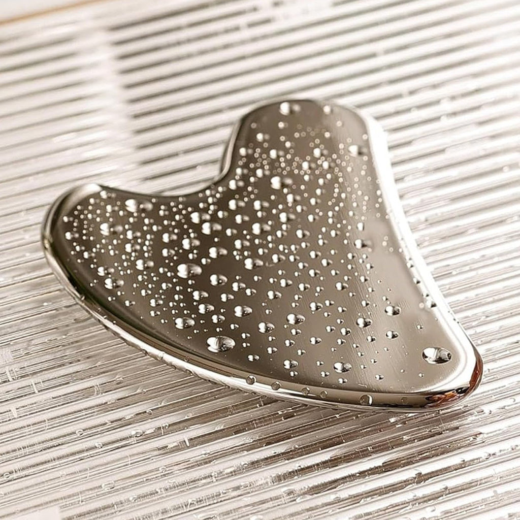 Heart-shaped stainless steel Gua Sha tool with water droplets, resting on a reflective, ridged surface.